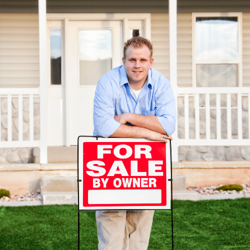man in blue shirt leaning on for sale by owner sign in front of a house